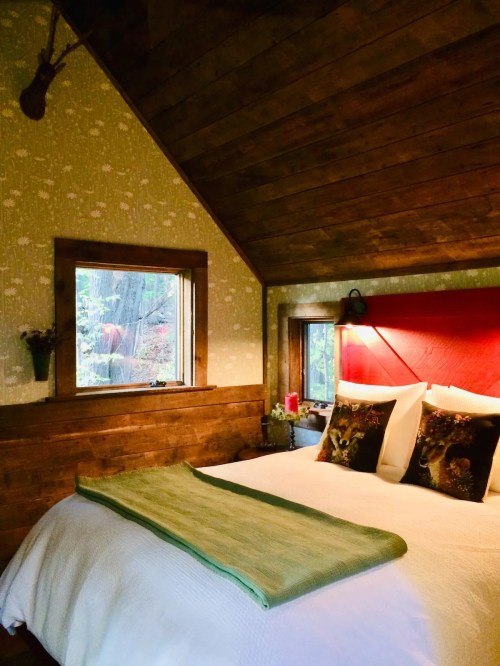 Cozy bedroom with wooden walls, a large bed, decorative pillows, and a window overlooking a forest.