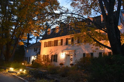A charming white house illuminated by warm light, surrounded by autumn trees at dusk.