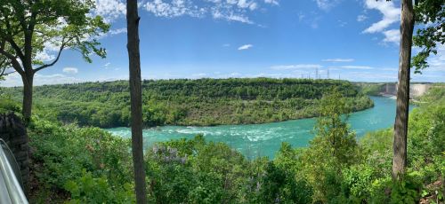 A panoramic view of a river winding through lush green hills under a bright blue sky with scattered clouds.