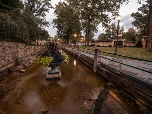 A serene park scene featuring a stone fountain surrounded by trees and a calm waterway, illuminated by soft evening light.