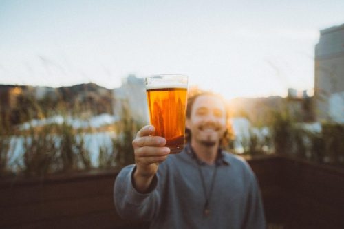 A person holds up a glass of amber beer, smiling against a sunset backdrop on a rooftop.