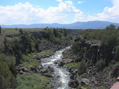 A winding river flows through a rocky canyon, surrounded by greenery and distant mountains under a blue sky.