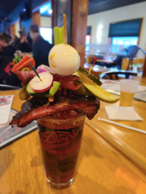 A tall, colorful Bloody Mary garnished with bacon, vegetables, and a quail egg, served in a glass on a wooden table.