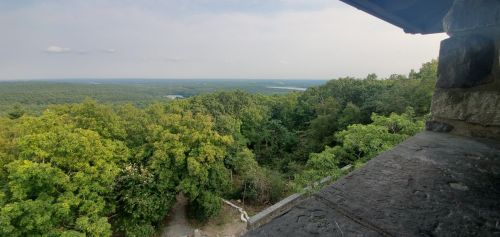 A panoramic view of lush green trees and distant hills under a cloudy sky, seen from a stone structure.