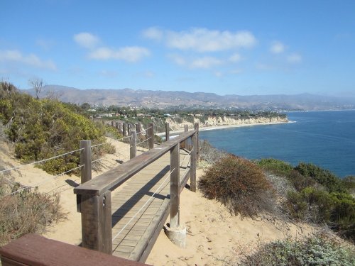 A wooden walkway along a sandy path overlooking a coastal landscape with cliffs and blue ocean under a clear sky.