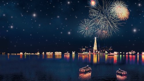 A festive night scene with fireworks over a decorated waterfront, featuring a glowing Christmas tree and colorful lights.