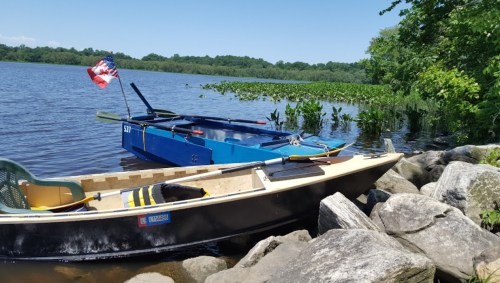 Two boats docked by a river, with one displaying an American flag, surrounded by greenery and rocky shoreline.
