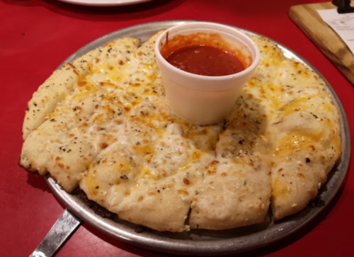 A round pizza with cheesy crust and a cup of marinara sauce in the center, served on a metal tray.