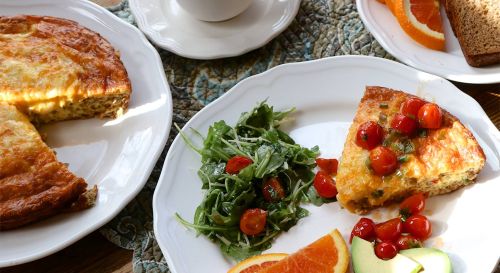 A plate with a slice of frittata topped with tomatoes, arugula salad, orange slices, and a piece of bread.