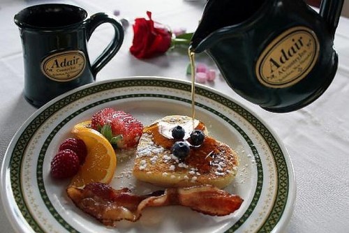 A plate of heart-shaped pancakes topped with berries and syrup, accompanied by bacon and a rose, with mugs in the background.