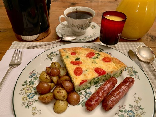 A breakfast plate with frittata, roasted potatoes, sausages, coffee, and juice on a wooden table.