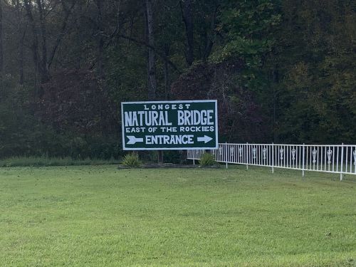 Sign for the longest natural bridge east of the Rockies, with arrows pointing to the entrance, surrounded by greenery.