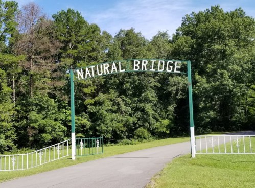 A green archway sign reading "NATURAL BRIDGE" at the entrance to a wooded area with a paved path.