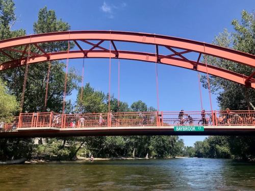 A red arch bridge spans a river, with people walking on it and trees lining the banks under a clear blue sky.