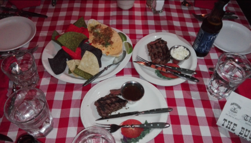 A table set with a checkered tablecloth, featuring plates of steak, chips, and a drink, surrounded by glasses and utensils.