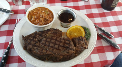 A plate with grilled steak, baked beans, a slice of orange, and a small cup of sauce, on a red and white checkered tablecloth.