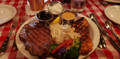 A plate of grilled steak with mashed potatoes, broccoli, grilled vegetables, and dipping sauces on a checkered tablecloth.