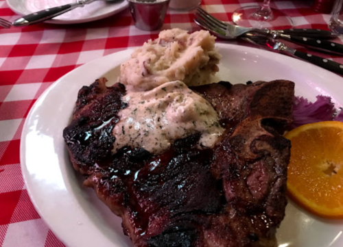 A large grilled steak topped with creamy sauce, served with mashed potatoes and an orange slice on a checkered tablecloth.