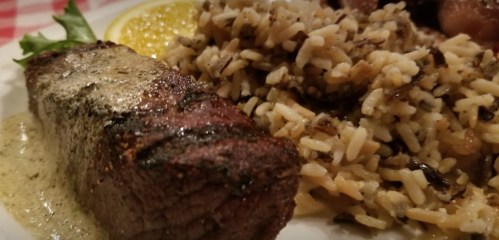 A plate featuring grilled steak with a creamy sauce, served alongside seasoned rice and a slice of lemon.