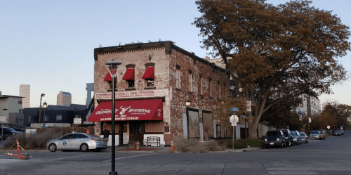 Historic brick building with red awnings, street lamps, and trees, set against a city skyline in the background.