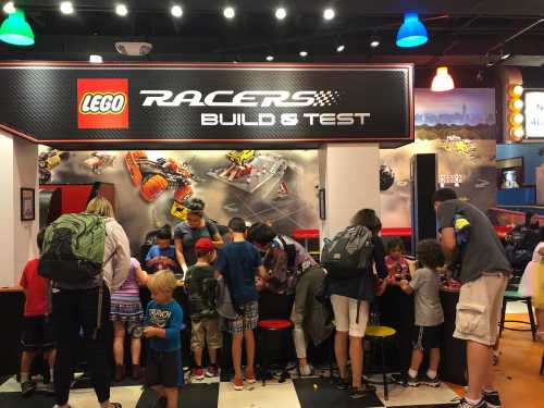 A group of children and adults gather around a LEGO Racers Build & Test station, engaging with various LEGO sets.