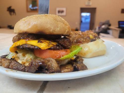 A close-up of a juicy burger with cheese, lettuce, and tomato on a plate, alongside a side of meat.