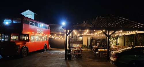 A red double-decker bus parked outside a cozy restaurant with string lights and outdoor seating at night.