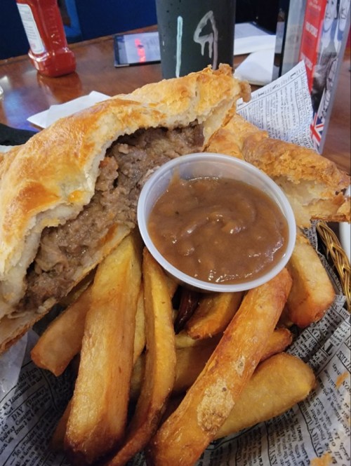 A hearty meat pie with a side of golden fries and a small cup of brown gravy on a newspaper-lined plate.