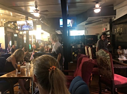 A lively bar scene with patrons seated at tables, watching TV, and a bartender serving drinks in a cozy atmosphere.