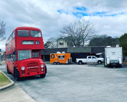 A red double-decker bus parked in a lot with food trucks and a pickup truck in the background under a cloudy sky.