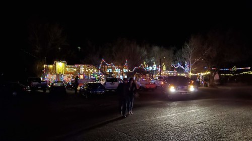 A festive scene at night with colorful holiday lights on buildings and trees, and people walking in the foreground.