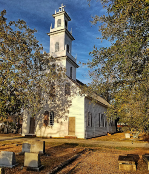 A historic white church with a tall steeple, surrounded by trees and a cemetery, under a clear blue sky.