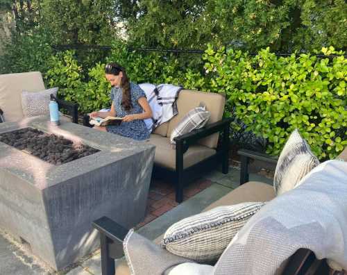 A woman in a blue dress sits on a patio, reading a book near a fire pit surrounded by greenery.