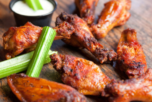 A plate of crispy chicken wings with celery sticks and a small cup of dipping sauce on a wooden surface.