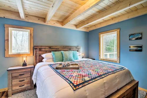 Cozy bedroom with a wooden bed, colorful quilt, and large windows, featuring a rustic design and soft lighting.