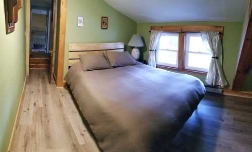 Cozy bedroom with a double bed, wooden accents, and green walls, featuring large windows and a lamp.