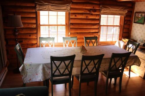 A rustic dining room with a long table and eight chairs, surrounded by wooden walls and bright windows.