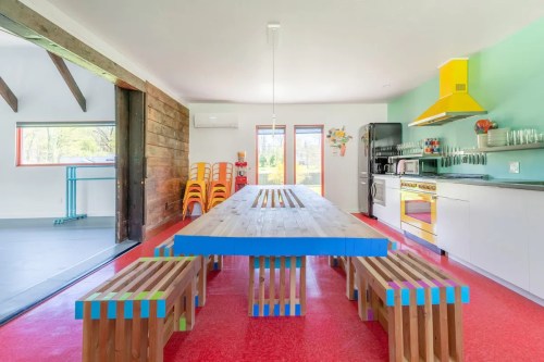 Bright, modern kitchen with a large wooden table, colorful benches, and a yellow hood over the stove. Red flooring.