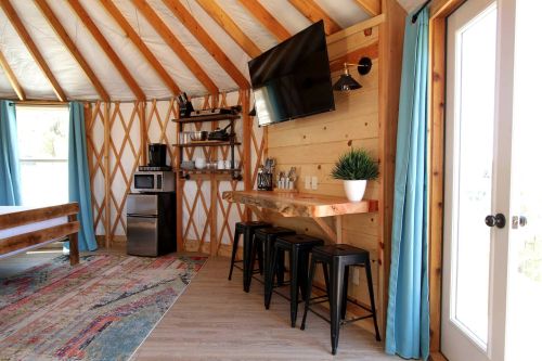 Cozy interior of a yurt with wooden beams, a TV, kitchenette, and seating area featuring a rug and blue curtains.