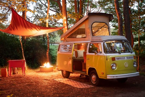 A vintage yellow VW camper van parked in a forest, with a fire pit and a colorful tarp overhead. Cozy camping scene.