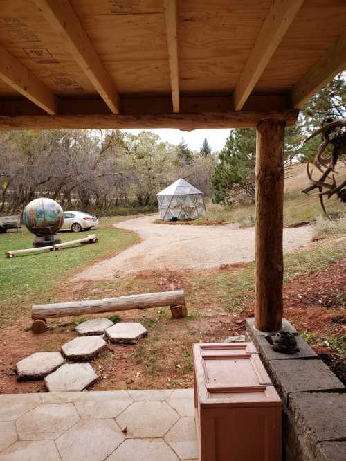 View from a porch showing a gravel path, a geodesic dome, a globe sculpture, and a car in a natural setting.