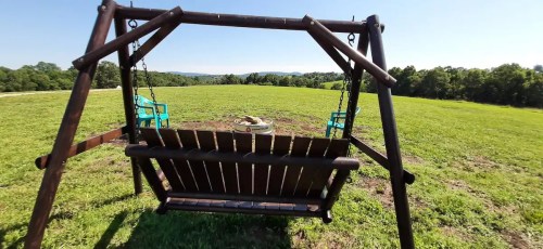 A wooden swing set in a grassy field, with two blue chairs and a scenic view of distant hills under a clear sky.