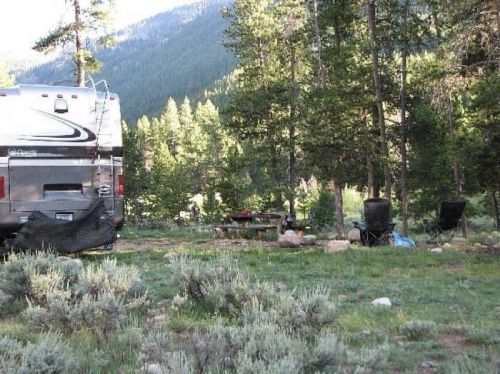 A campsite with an RV, picnic table, and chairs surrounded by trees and mountains in the background.