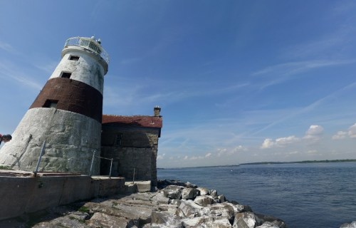 A lighthouse stands on rocky shore under a blue sky with scattered clouds, overlooking a calm body of water.