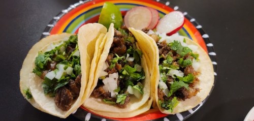 Three tacos filled with meat, topped with onions and cilantro, served with lime and radishes on a colorful plate.