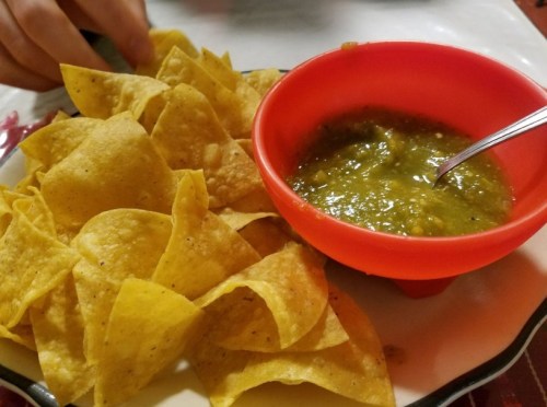 A plate of golden tortilla chips next to a small red bowl of green salsa.