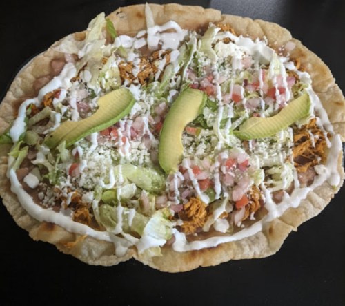A flat tortilla topped with shredded chicken, lettuce, tomatoes, avocado slices, cheese, and drizzled with sour cream.