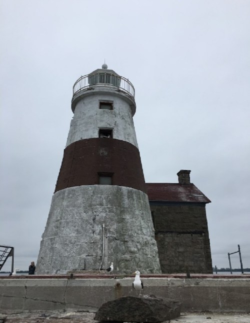 A tall, weathered lighthouse with a gray and white exterior, surrounded by a rocky shoreline under a cloudy sky.