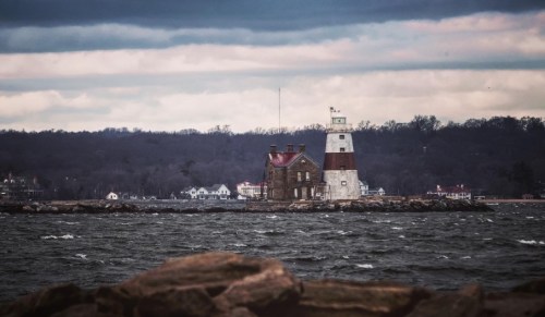 A lighthouse stands on a rocky shore, surrounded by choppy waters under a cloudy sky. Trees line the distant shore.
