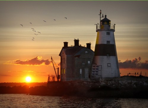 A lighthouse stands by the shore at sunset, with birds flying in the sky and a warm glow reflecting on the water.